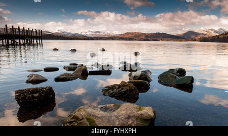 La costa rocciosa del lago Windermere nel Lake District inglese, con un molo e montagne offrono lo sfondo. Foto Stock