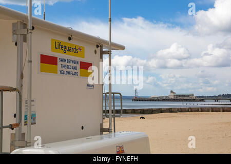 Nuotare tra il giallo e il rosso bandiere segno su RNLI bagnini chiosco in spiaggia con molo di Bournemouth in distanza Foto Stock