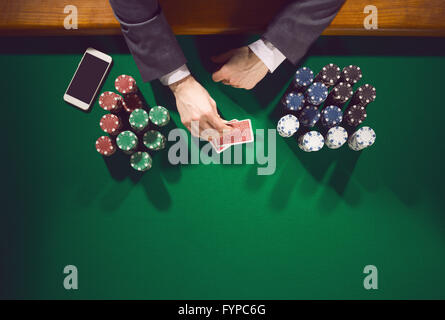 Maschio elegante poker player con lo smartphone guardando le sue carte con pile di chips in tutto Foto Stock