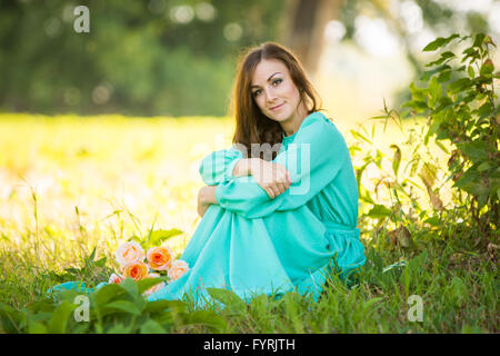 Una bella ragazza seduta sotto l'ombra degli alberi, giace ai piedi del suo bouquet di rose Foto Stock