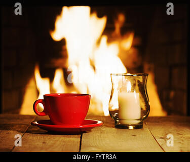 Red tazza di caffè o di tè e la candela sul tavolo di legno vicino al camino. Inverno e vacanze di Natale concetto. Foto Stock