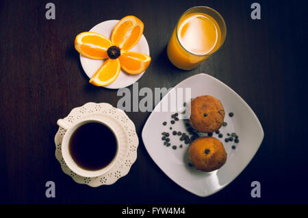 La colazione sul legno scuro superficie comprendente caffè e muffin, succo d'arancia e fette di arance posto sulla piccola piastra bianca Foto Stock