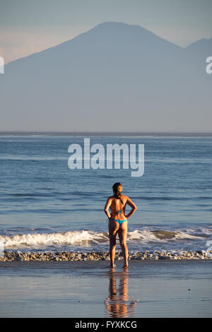 Una donna, nelle prime ore del mattino, in piedi sulla riva che guarda al mare nella baia di Jimbaran, Bali, con montagne in lontananza. Foto Stock