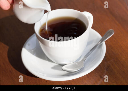 Versare il latte in una tazza di caffè nero su un tavolo di legno. Foto Stock