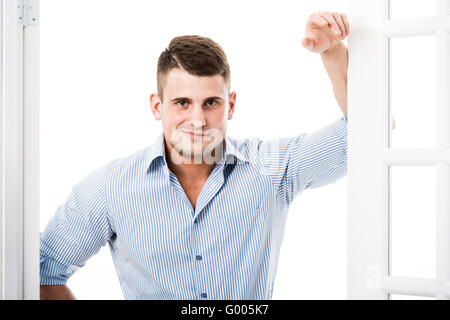 Ritratto di giovane casual uomo appoggiato contro il telaio di una porta aperta su sfondo chiaro sorridente e guardando la fotocamera con la mano in tasca Foto Stock