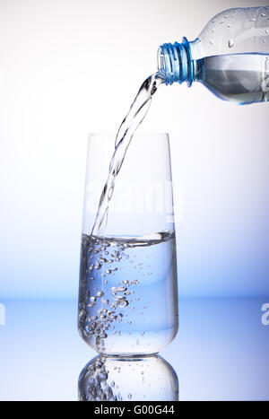 Versare acqua dalla bottiglia di plastica in half-filled bicchiere con la riflessione su bianco e lo sfondo blu Foto Stock