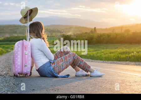 Lonely ragazza seduta sulla strada vicino alla sua valigia Foto Stock