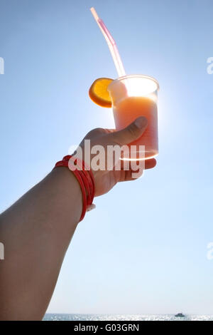 Mano che tiene un cocktail tequila sunrise sullo sfondo del mare con yacht di lusso all'orizzonte Foto Stock