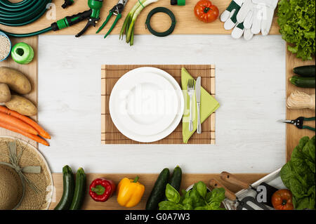 Utensili da giardinaggio e verdure fresche che compongono un telaio, tabella imposta al centro con lavastoviglie, forchetta e coltello, vista dall'alto Foto Stock