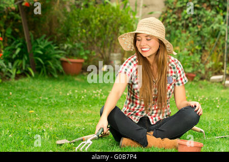 Giovane donna seduta su erba usando utensili da giardinaggio mentre si lavora all'aperto Foto Stock