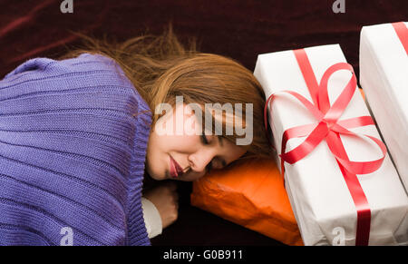 Brunette coperto con indumenti di colore viola dorme, appoggiato la testa tra presenta, carta di avvolgimento bianca e nastro rosso Foto Stock