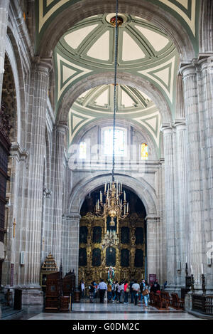 Navata interna della Cattedrale Metropolitana di Città del Messico con persone | CITTÀ DEL MESSICO, Messico — Una navata laterale della Catedral Metropolitana si estende verso un retablo barocco dorato sotto una volta a costoloni in pietra chiara di chiluca, con visitatori riuniti intorno all’altare sottostante. Le 40 colonne in cantera della cattedrale sostengono 51 volte su cinque navate, un sistema strutturale ridotto rispetto a un progetto originale a sette navate di Siviglia specificamente perché i terreni argillosi dell’ex Lago di Texcoco non potevano sopportare un carico maggiore. Foto Stock