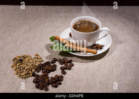 Pila di due tipi diversi di java bean e erbe aromatiche accanto fumante tazzina piena di caffè e piattino sulla tovaglia grigio Foto Stock