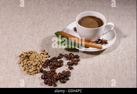 Pila di due tipi di colore diverso i chicchi di caffè accanto a pieno tazzina e piattino sulla tovaglia grigio Foto Stock