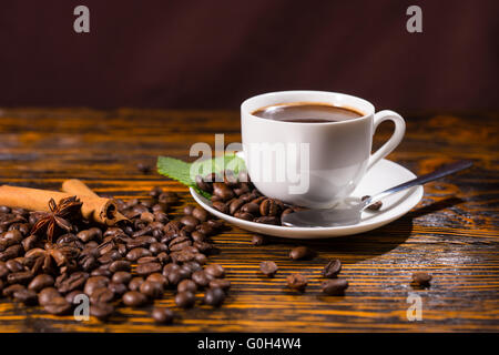 Close up ancora in vita della tazza bianca con piattino contenenti caffe' macinato di fresco in appoggio sulla tavola in legno rustico Top sparse con chicchi di caffè tostati, bastoncini di cannella e anice stellato. Foto Stock