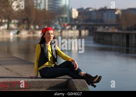 Bella donna asiatica in maglione giallo e rosso con velo seduti su un fiume in una città europea Foto Stock