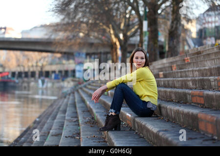 Bella donna asiatica in felpa gialla seduti su un fiume in una città europea Foto Stock