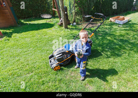 Carino, Little Boy mows prato. boy ispeziona la falciatrice, prima di guidare o di sinistra dell'olio motore. Foto Stock