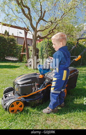 Carino, Little Boy mows prato. boy ispeziona la falciatrice, prima di guidare o di sinistra dell'olio motore. Foto Stock