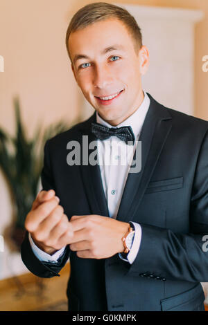 Elegante moda giovane uomo in nero Tuxedo regolando i suoi gemelli mentre guardando la telecamera in ambienti interni Foto Stock