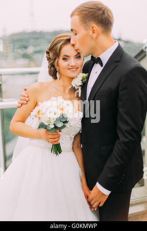 Felice bella giovane, lo sposo baciare sposa la fronte sulla terrazza con il paesaggio sullo sfondo Foto Stock