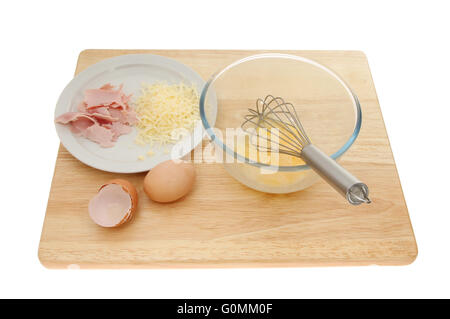 Ingredienti frittata, uova, formaggio e prosciutto con una frusta e recipiente di miscelazione su un tagliere di legno isolata contro bianco Foto Stock