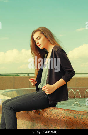 Ragazza solitaria vuole qualcuno a parlare. Indossando grigio chiaro maglione, pantaloni neri, un youg american donna seduta su granito rosso fontana, triste e depresso, perso nel pensiero. Effetto Instagram Foto Stock