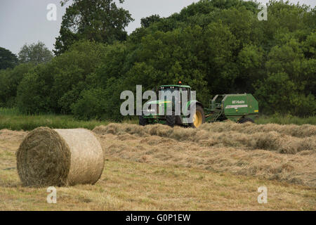 Di fieno o insilato, grande Ouseburn, North Yorkshire, Inghilterra - Balla rotonda in campo con trattore verde & guida al di là di lavoro, tirando un'imballatrice. Foto Stock