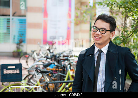 Giovani asiatici sorridente uomo d affari o studente in piedi nella parte anteriore della bicicletta rack parcheggio su strada di città, con spazio di copia Foto Stock