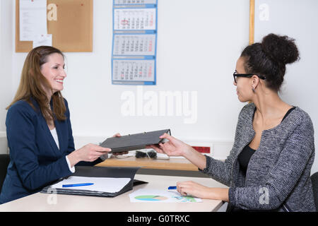 Due donne di business sono seduti ad un tavolo in ufficio. Una donna è la consegna di un file per l'altra donna. Foto Stock