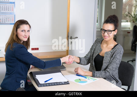 Due donne di business stanno facendo una stretta di mano durante una riunione. Essi sono qui seduti a un tavolo in ufficio. Entrambe le donne stanno cercando di Foto Stock