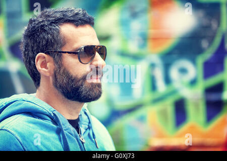 Closeup ritratto di uomo bello elegante con barba e occhiali da sole, stando in piedi sopra colorata città sullo sfondo della parete Foto Stock