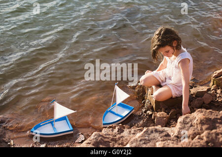 Ragazza seduta sulle rocce dal lago di giocare con barche giocattolo Foto Stock