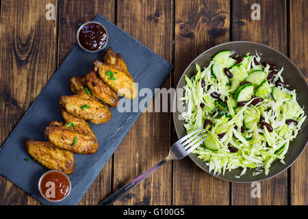 Ali di pollo su una piastra di ardesia e insalata su sfondo di legno Foto Stock