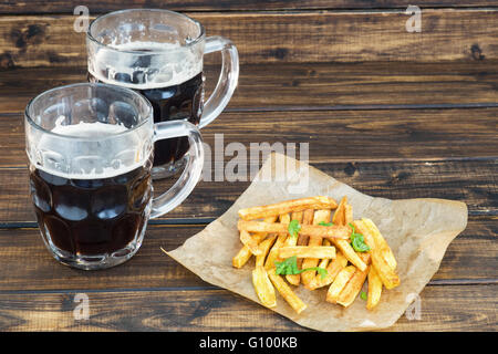 Due boccali di birra scura con patate fritte su sfondo di legno Foto Stock