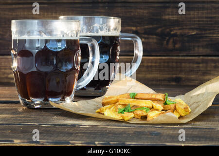 Due boccali di birra scura con patate fritte su sfondo di legno Foto Stock