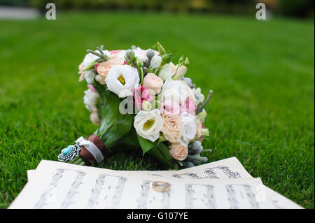 Due anelli di nozze su un foglio di carta con note musicali. Bouquet nozze sull'erba verde. Foto Stock