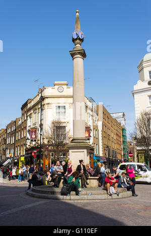 I londinesi godendo il sole seduti sotto la meridiana pilastro di Seven Dials, un piccolo nodo stradale in Covent Garden di Londra Foto Stock