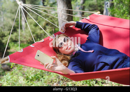Giovane ragazza sorridente in bicchieri godere in rosso amaca in foresta. Redhead donna con lentiggini sorrisi, rende selfie con lo smartphone. Foto Stock