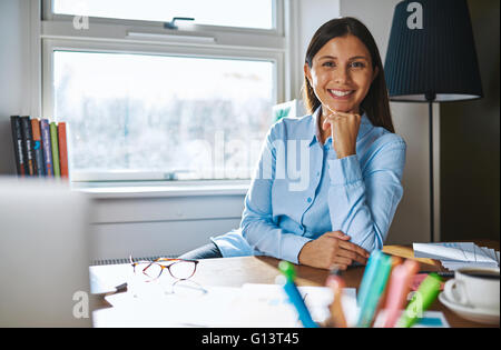 Donna sorridente con la mano sul mento indossando maglietta blu mentre si è seduti alla scrivania, ricoperti con carte, caffè e occhiali da vista e finestra ho Foto Stock