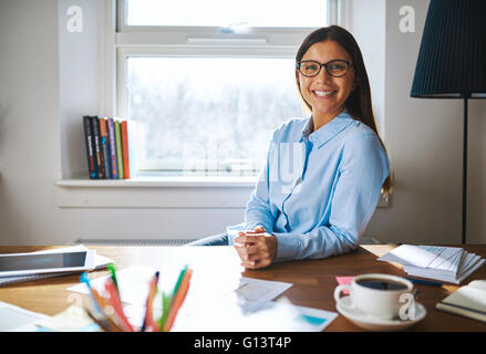 Lavoratori autonomi donna che indossa gli occhiali e maglietta blu alla scrivania con le mani giunte accanto alla tazza piena di caffè e giornali in home offic Foto Stock