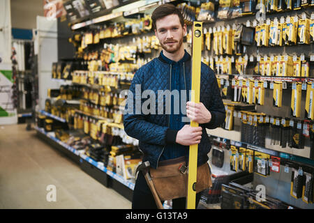 Giovane falegname tenendo un livello di costruttori in un negozio di ferramenta in piedi nella corsia a fianco del merchandise sorridente in dotazione Foto Stock