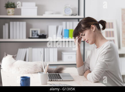 La donna a casa, seduti alla scrivania e lavorare con un computer portatile ha un mal di testa e toccando la fronte, un gatto sdraiato su Foto Stock