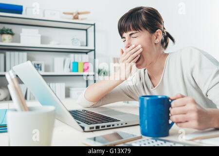 Stanco di sleepy donna sbadigli, lavorando alla scrivania in ufficio e in possesso di una tazza di caffè, superlavoro e la privazione del sonno concept Foto Stock