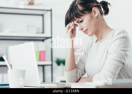 Triste stanco donna avente un brutto mal di testa, lei è seduta alla scrivania in ufficio e toccando il suo tempio Foto Stock