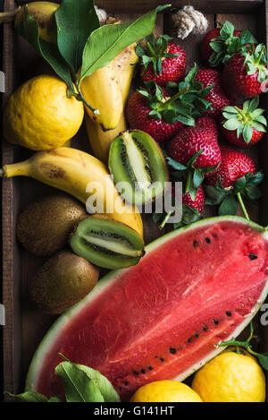Varie colorata frutta tropicale selezione nel vassoio di legno su sfondo scuro, vista dall'alto. Anguria, fragola, limoni e kiwi Foto Stock