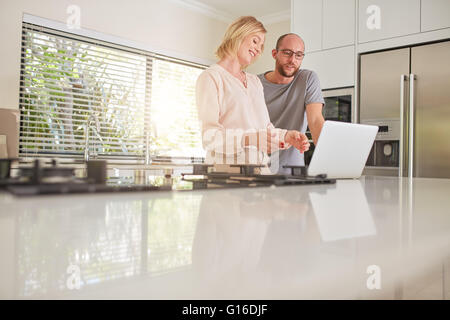 Tiro al coperto di una coppia felice utilizzando il computer portatile in cucina. La donna che mostra qualcosa sul laptop per l'uomo. Foto Stock