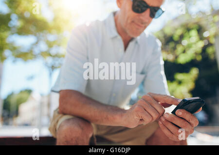 Close up ritratto di uomo anziano tramite telefono cellulare mentre è seduto su una panchina nella città in una giornata di sole. Focus su mani tenendo premuto MOB Foto Stock