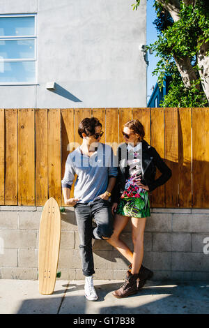 Coppia giovane in piedi insieme all'aperto, skateboard appoggiata sulla parete accanto all uomo Foto Stock