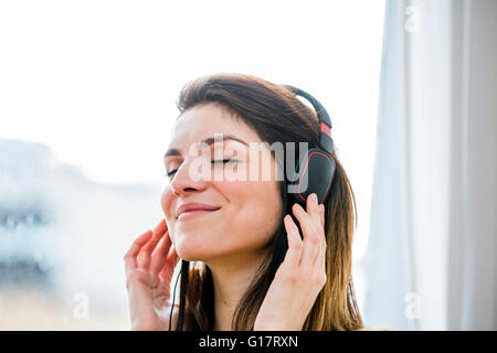 Bella giovane donna l'ascolto di musica per le cuffie nella parte anteriore della finestra appartamento Foto Stock